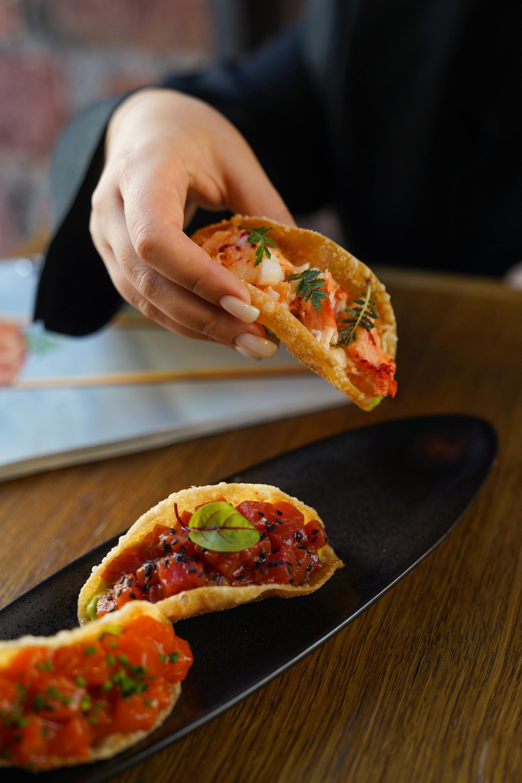 Close-up of a hand holding gourmet tacos on a black plate, showcasing intricate details.
